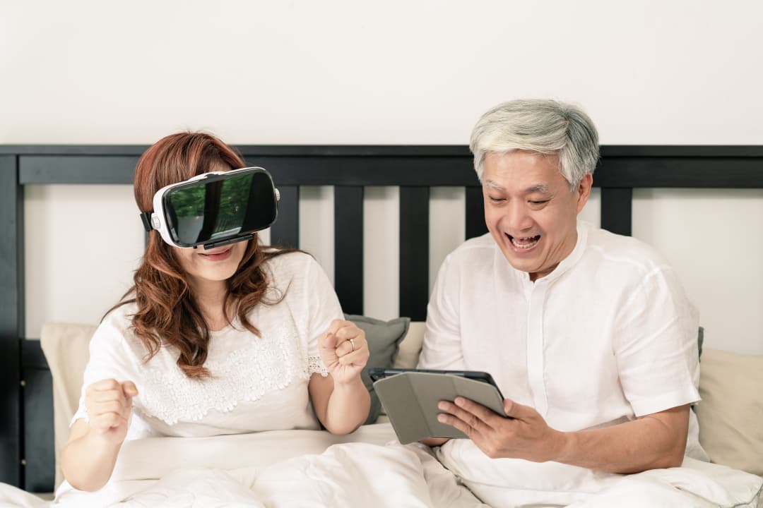 Et voksent par, der sidder i en senng. Kvinden har virtual reality-briller på, og manden holder en Ipad. De ser ud til have det sjovt.